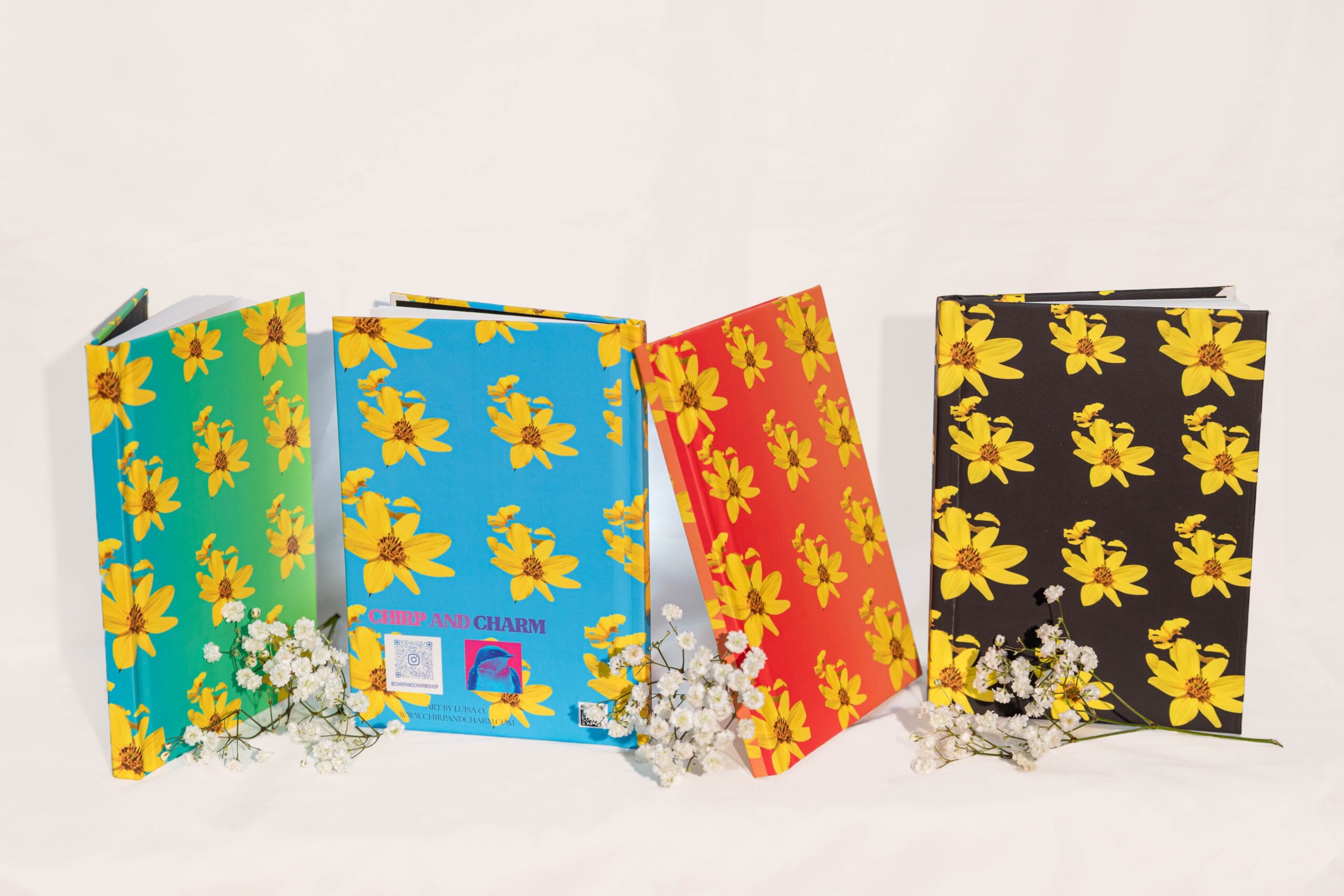 Four colorful notebooks with floral patterns, arranged in a row with small white flowers in front, on a white background.