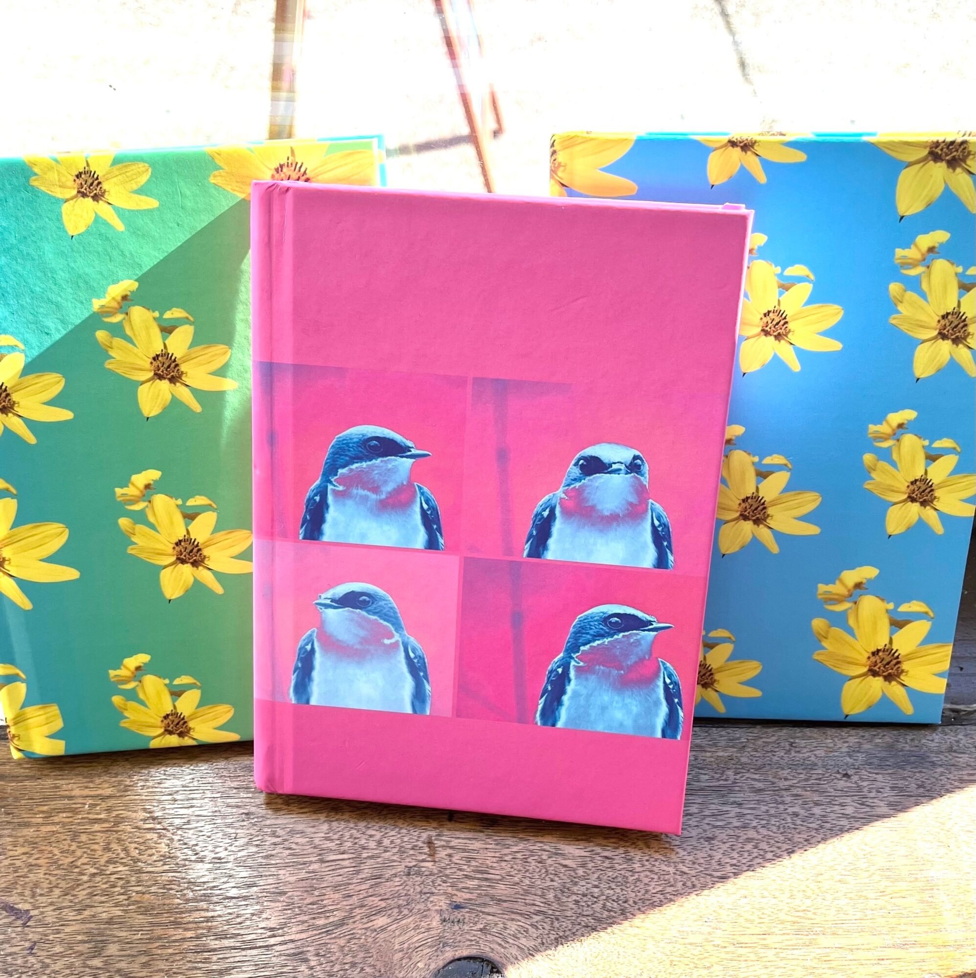 Pink notebook with four penguin images, flanked by colorful floral-patterned gift bags, on a textured surface outdoors.