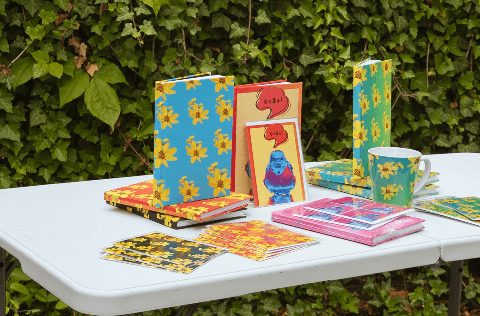 Table with colorful cards, a mug, and boxes outdoors against green leafy background.
