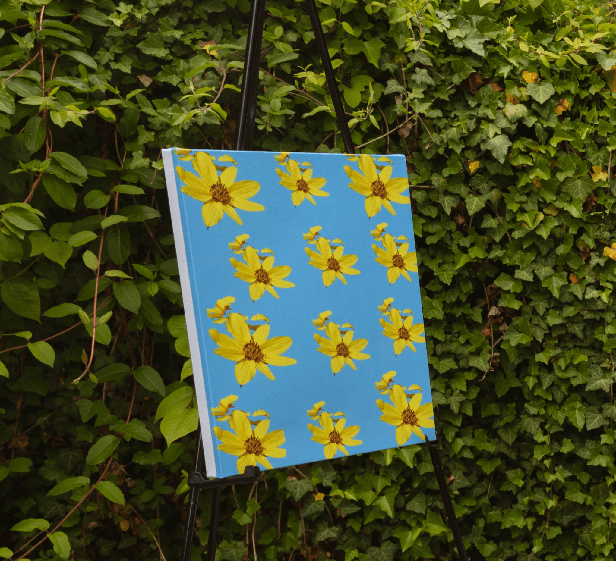 Painting with yellow flowers on a blue background, hanging outdoors among green foliage.