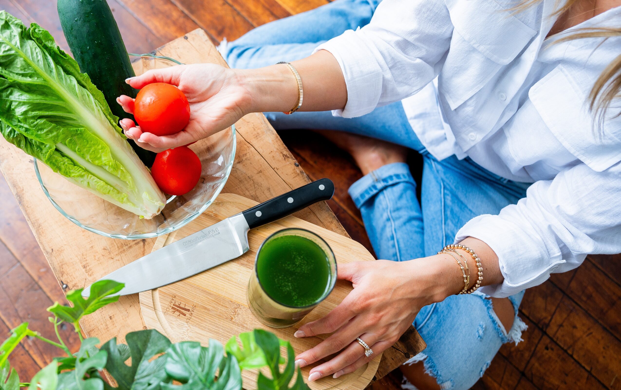 Person holding a tomato and a glass of green juice, sitting on a wooden floor with vegetables nearby.