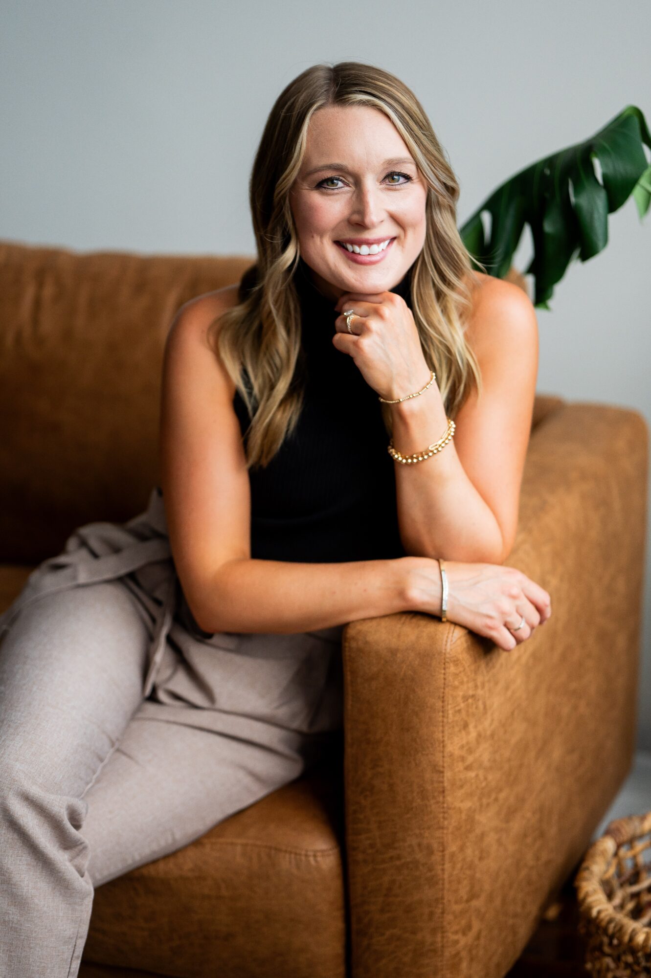 Woman with blonde hair smiling, sitting on a brown sofa, wearing a black top and beige pants, with a green plant in the background.