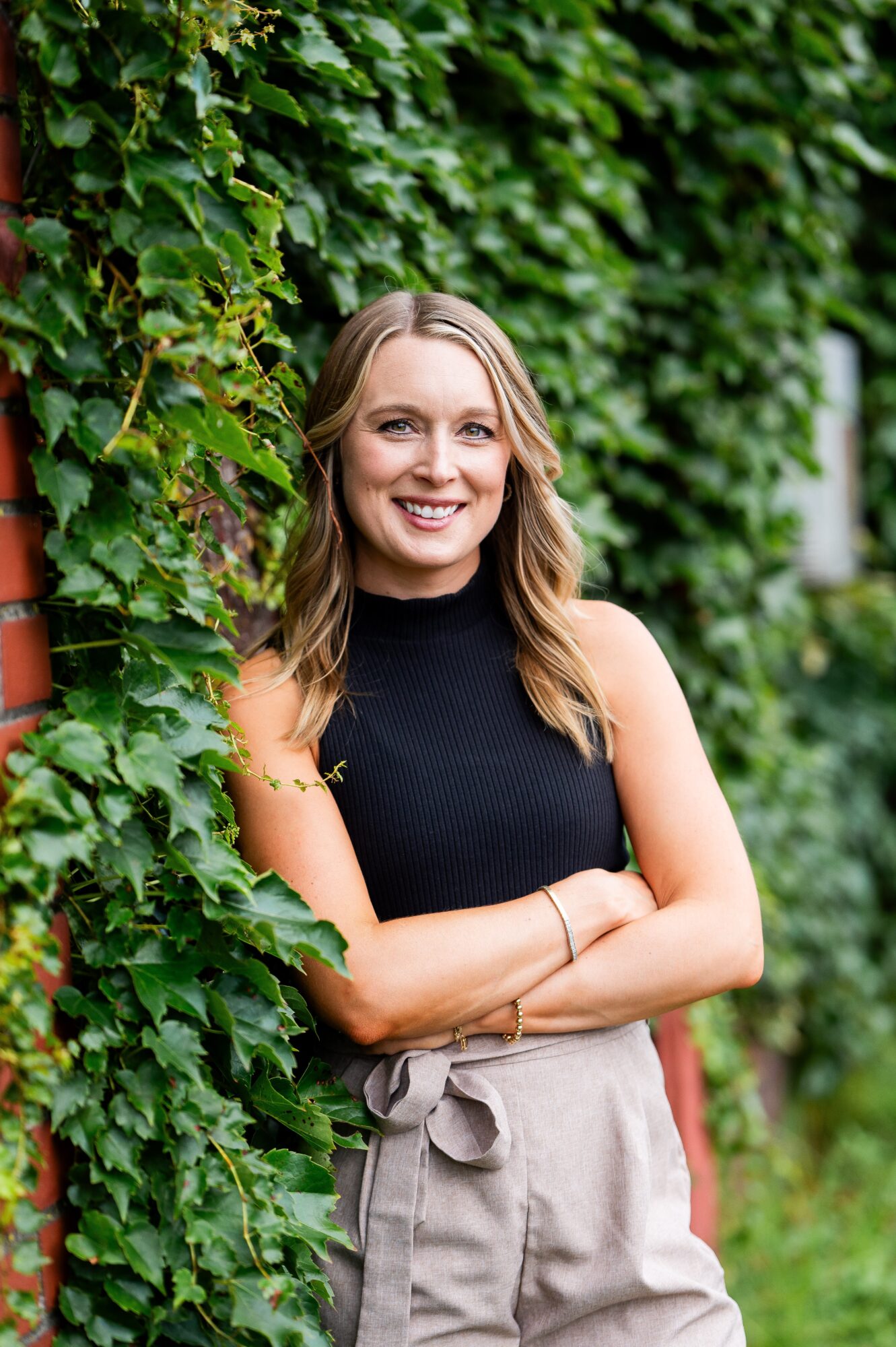 Smiling woman with long hair standing outdoors near green ivy-covered wall, arms crossed, wearing a black sleeveless top and beige pants.