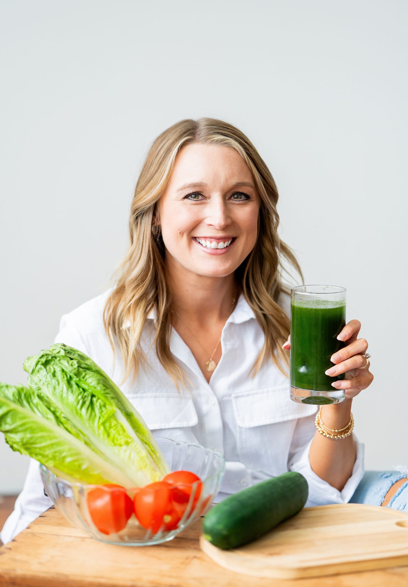 Smiling woman holding a glass of green juice, with lettuce, tomatoes, and a cucumber on the table.