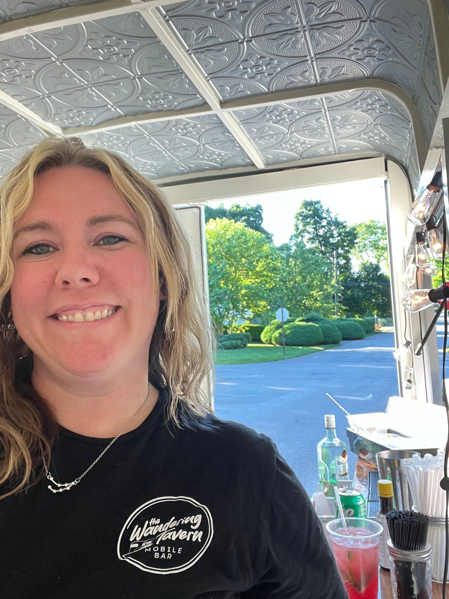 Smiling woman with blonde hair in a black shirt, sitting indoors near a window with a view of a pool and trees outside.