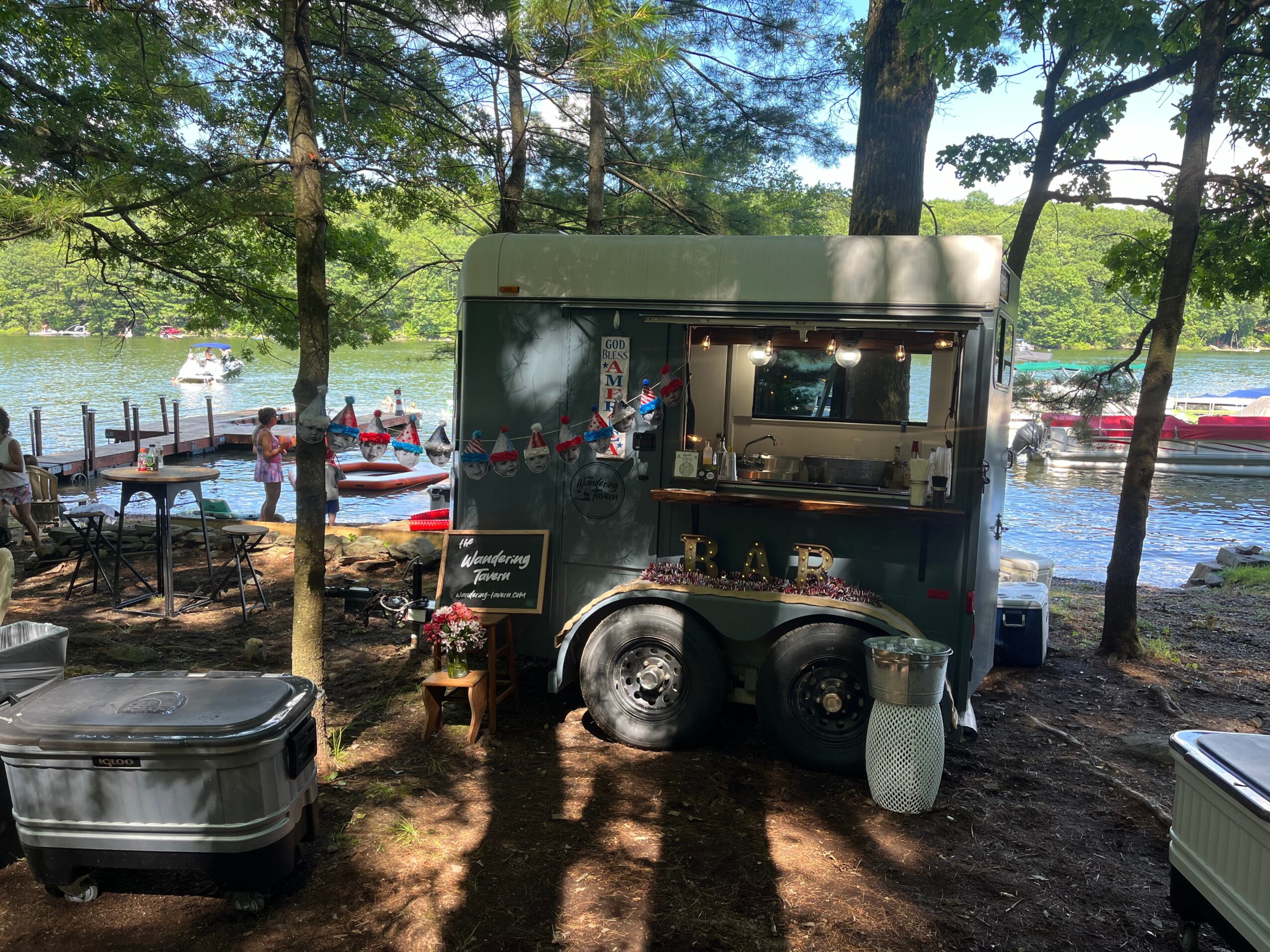 Food truck parked near a lake with trees, boats, and people in the background, under a sunny sky.