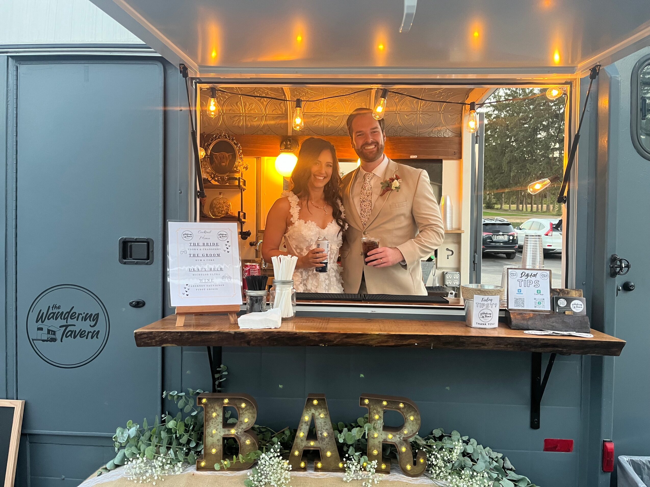 A couple stands inside a mobile bar with illuminated letters spelling 'BAR' and floral decorations, smiling at the camera.