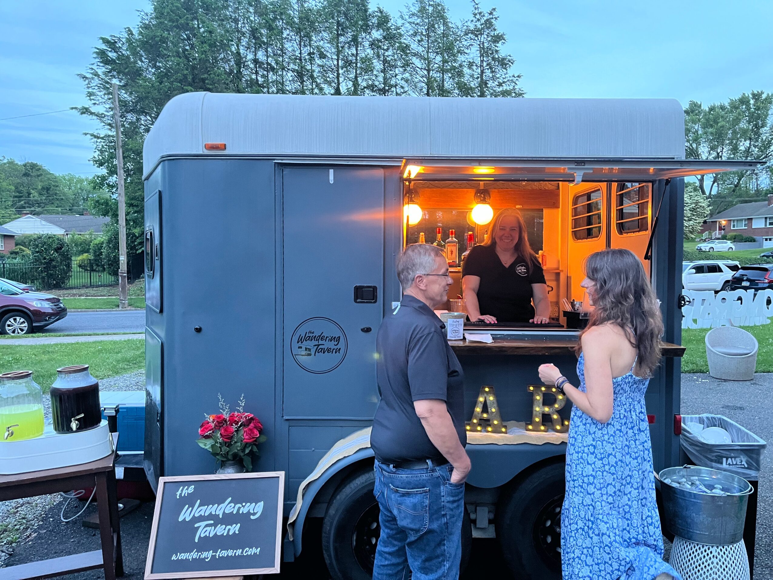 People at a food truck with a window serving customers outdoors, trees and cars in background, evening lighting.