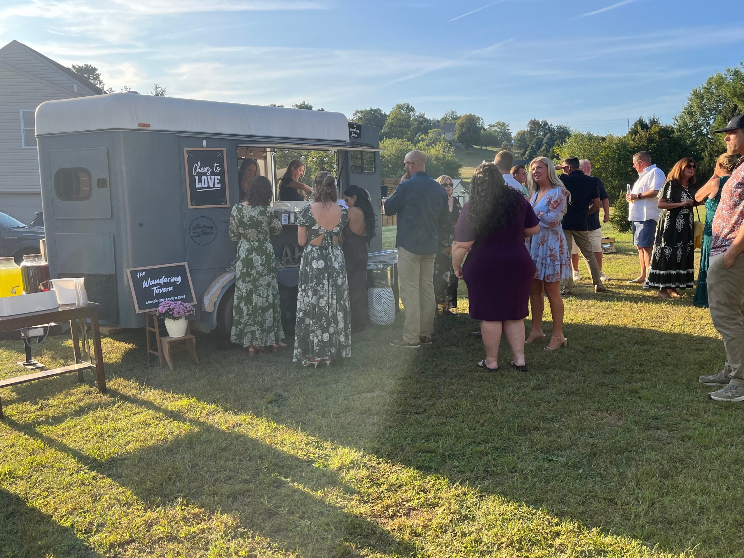 People gather around a food truck outdoors on a sunny day, with trees and a blue sky in the background.