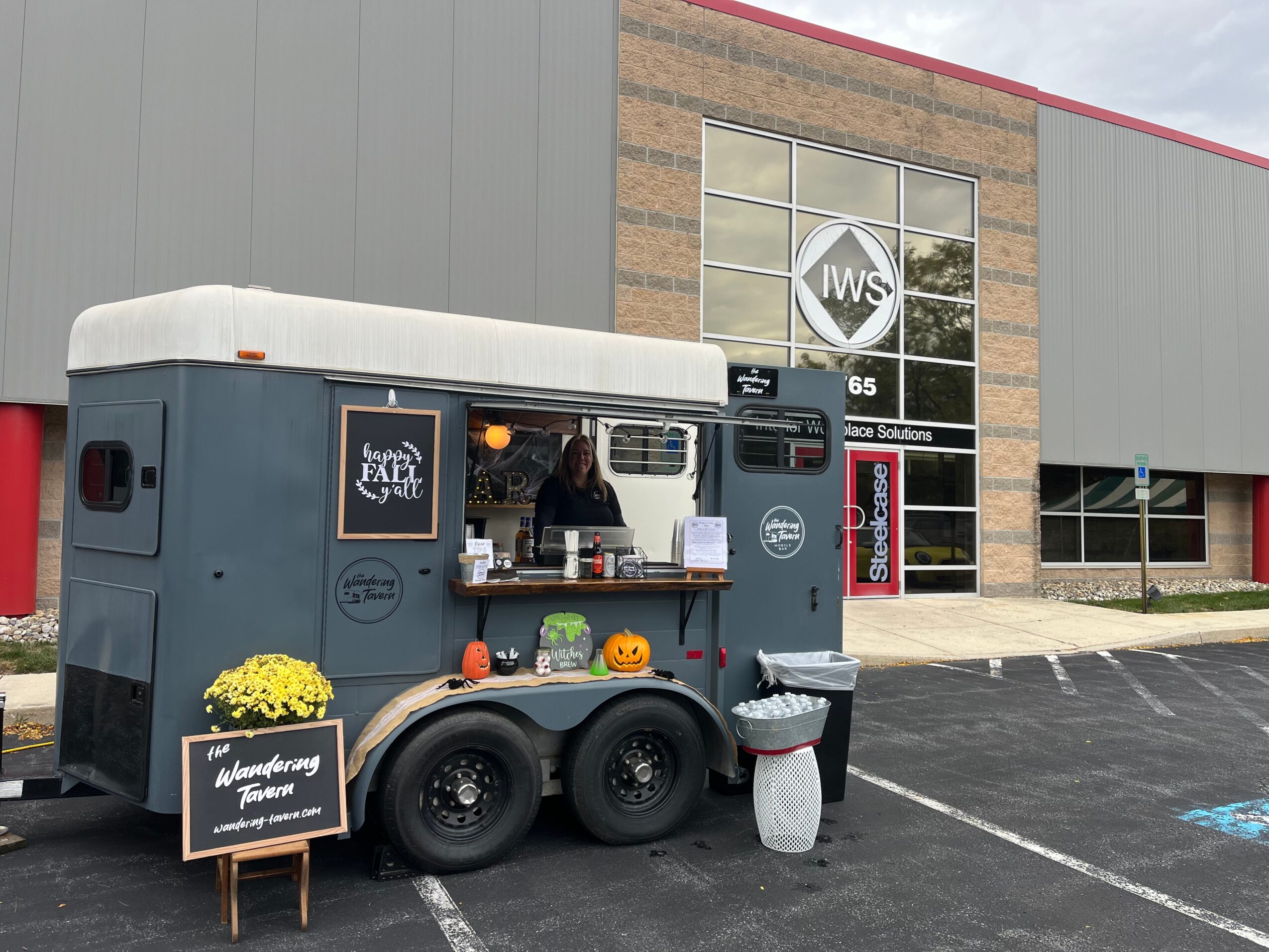 Food truck parked outside a building with large windows and a sign, with a chalkboard menu and decorations, in a parking lot.