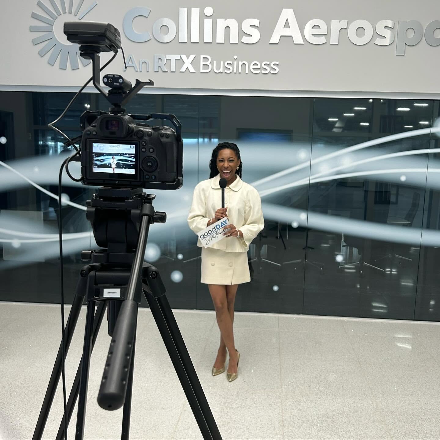 A woman stands in front of a camera at an event, with a backdrop displaying 'Collins Aerospace' and 'RTX Business'.