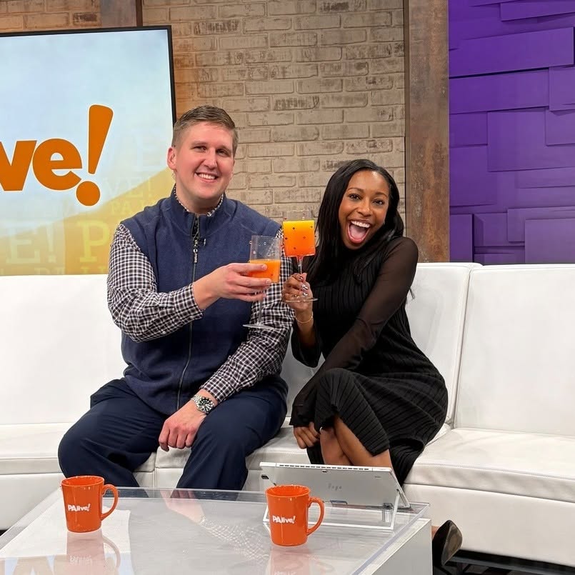 Two people sitting on a white couch, smiling and holding drinks, in a studio setting with a brick wall and purple panel background.