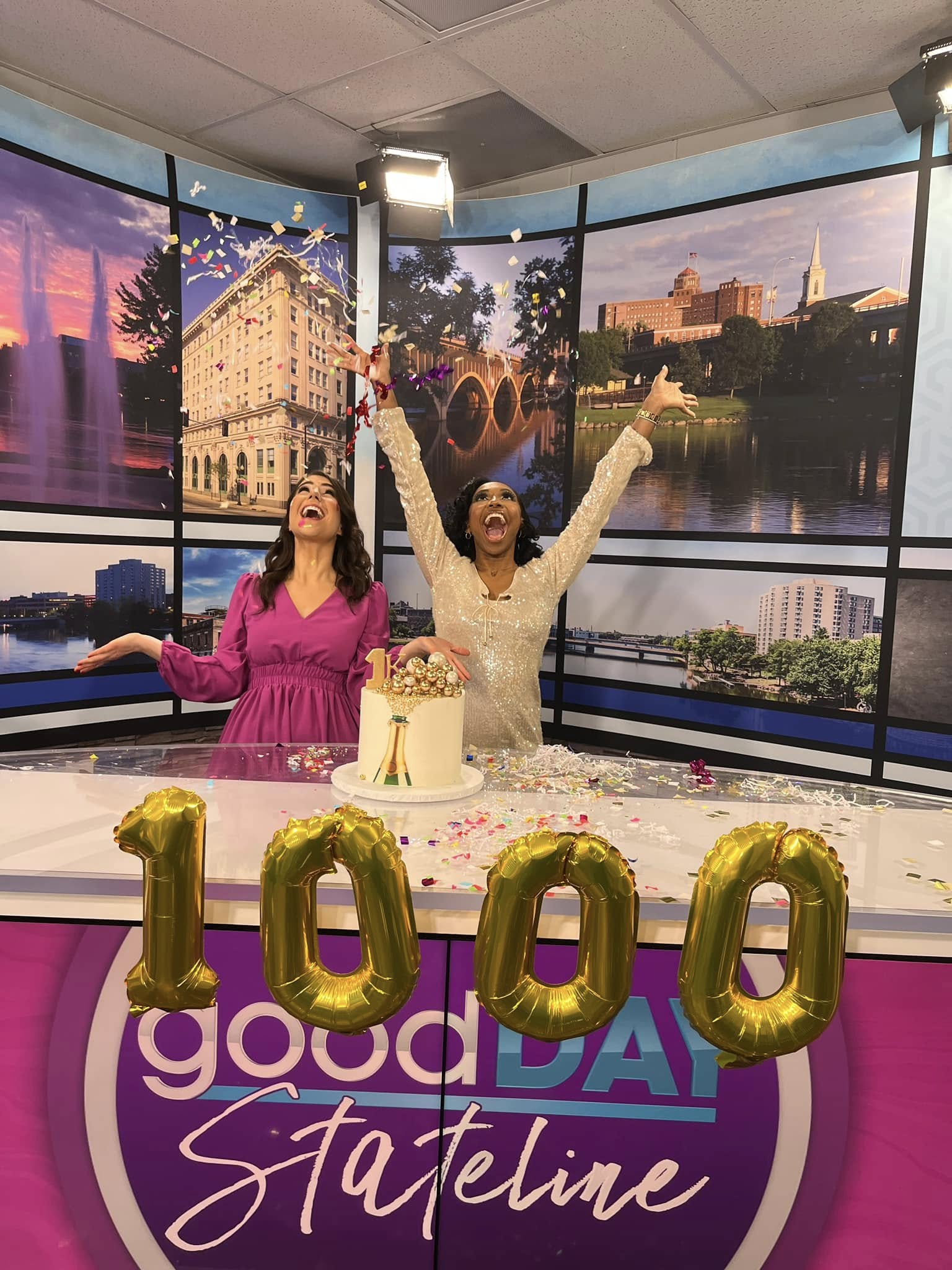 Two women celebrate a 100th birthday with balloons, cake, and a cityscape background, smiling and raising their arms.