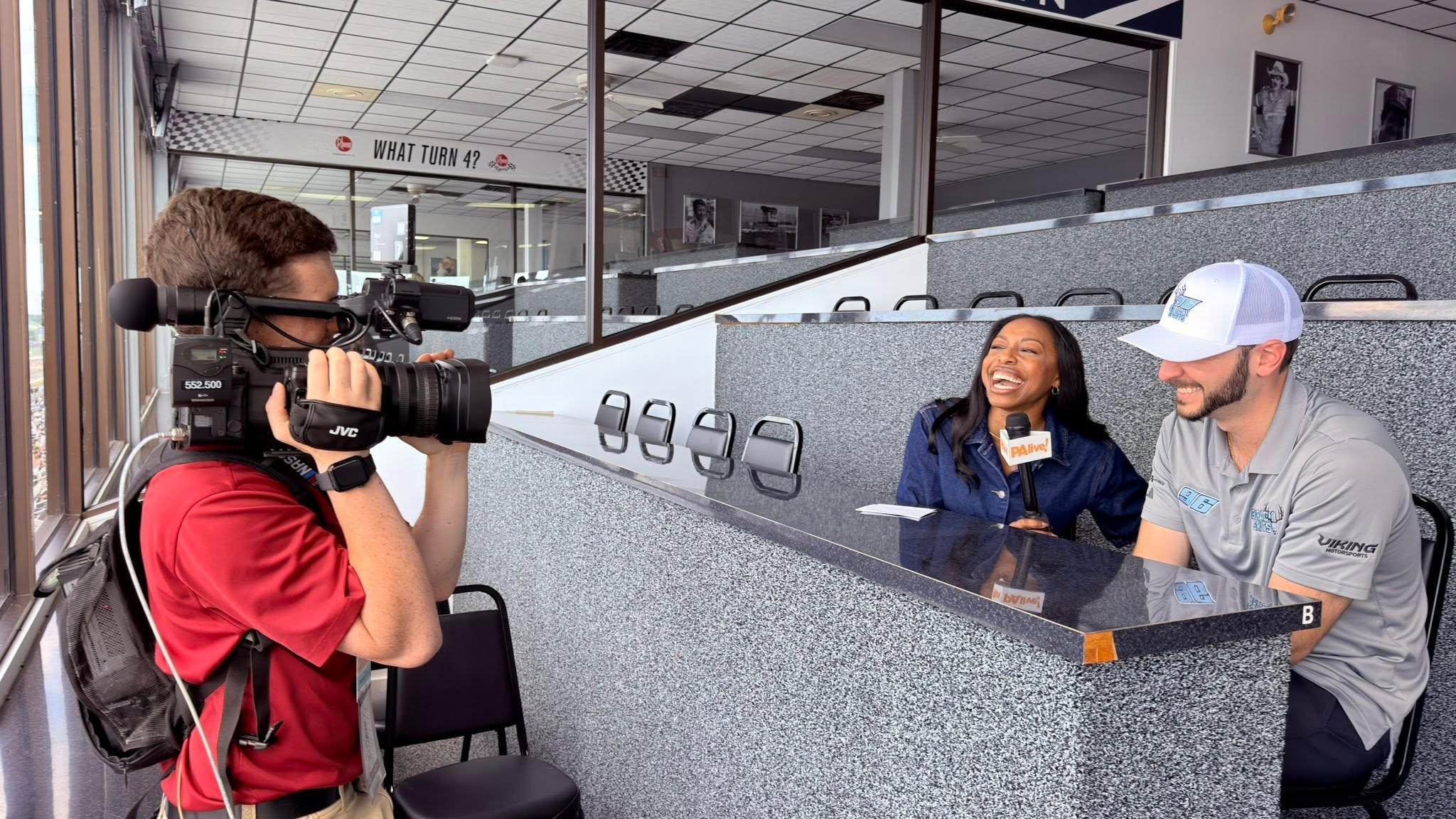 Person filming two people sitting at a counter in a public space, smiling and talking.