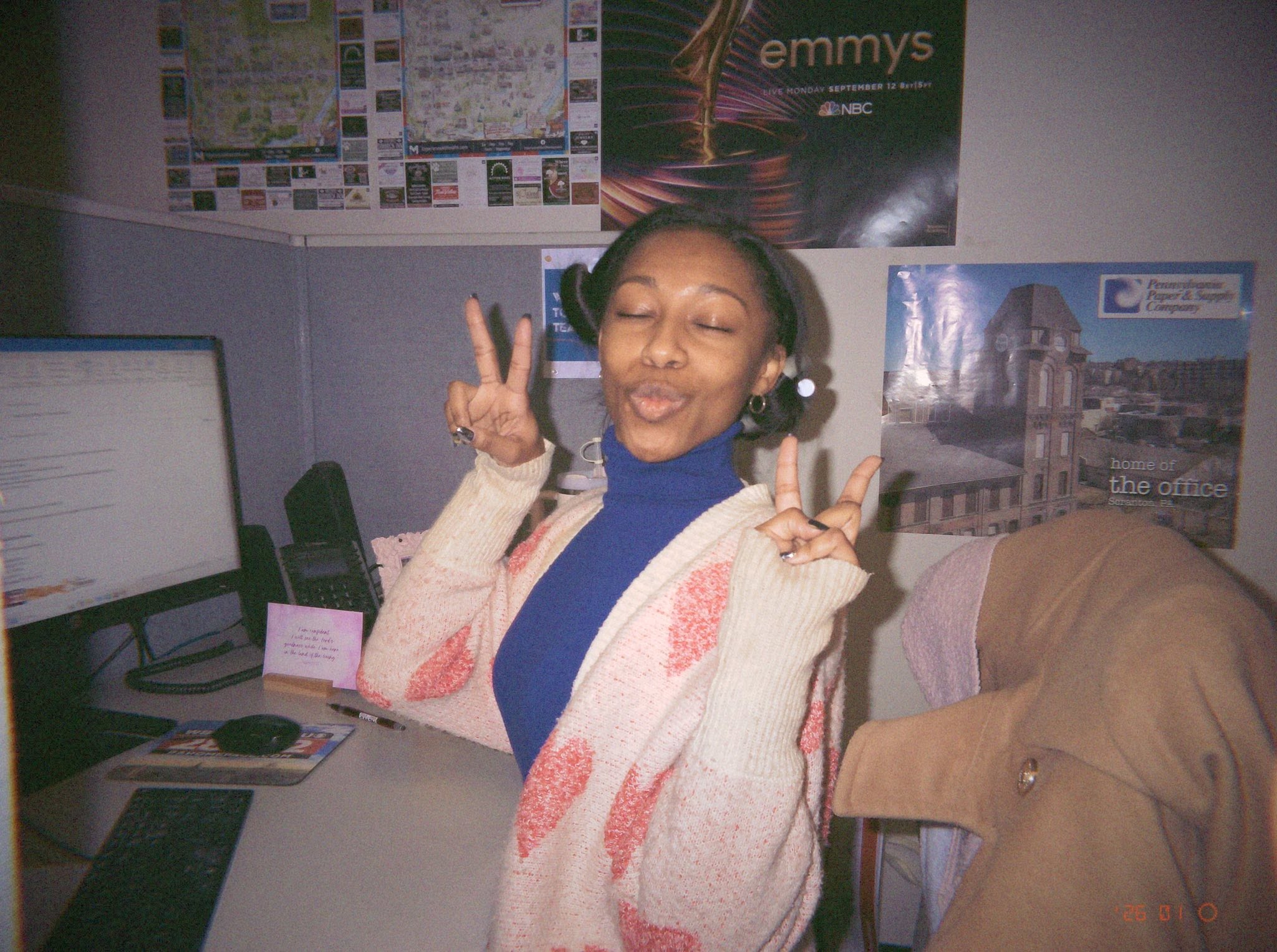Person with headphones smiling and making peace signs in an office, with computer and posters on the wall.