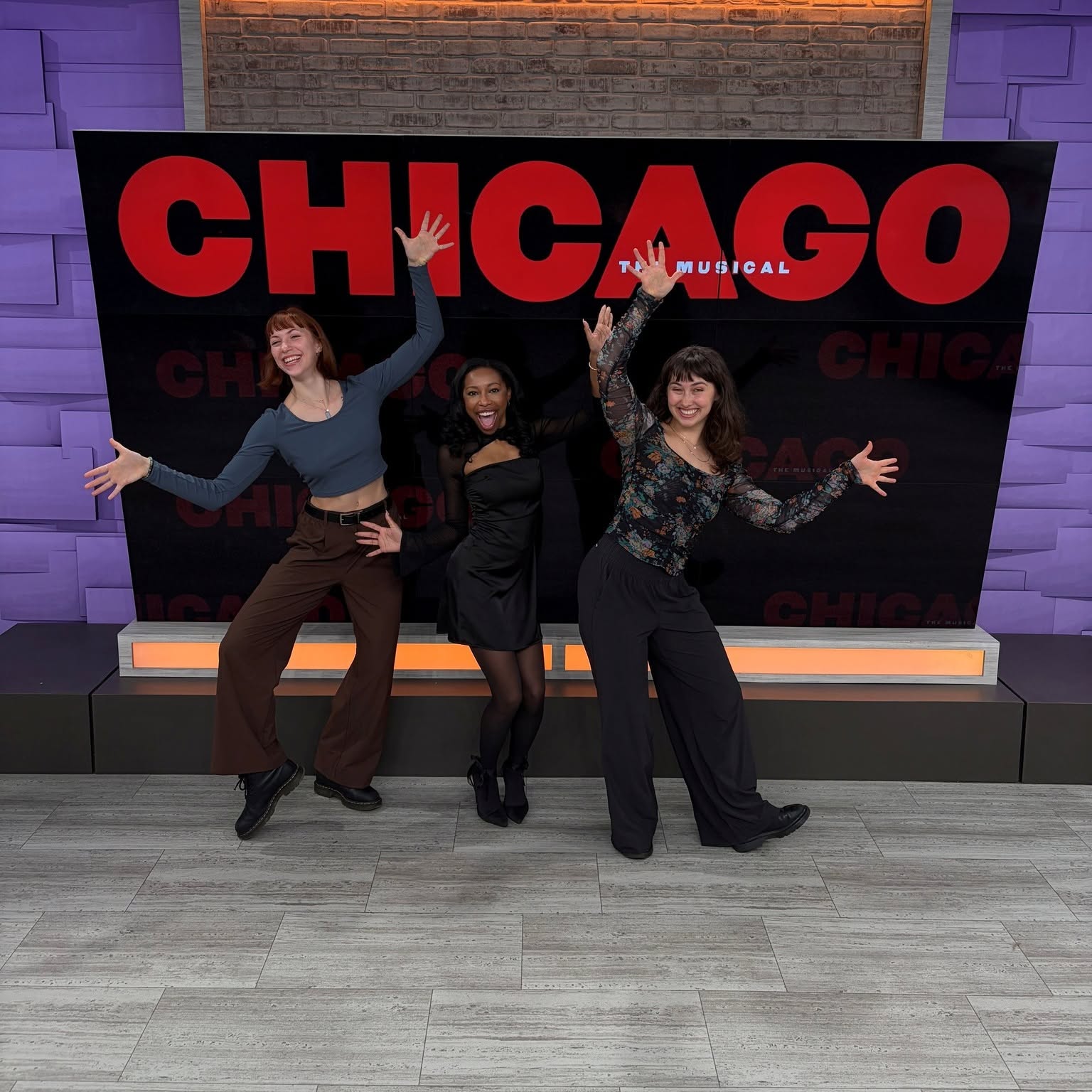Three women stand in front of a large red and black Chicago sign, smiling and posing with arms raised.