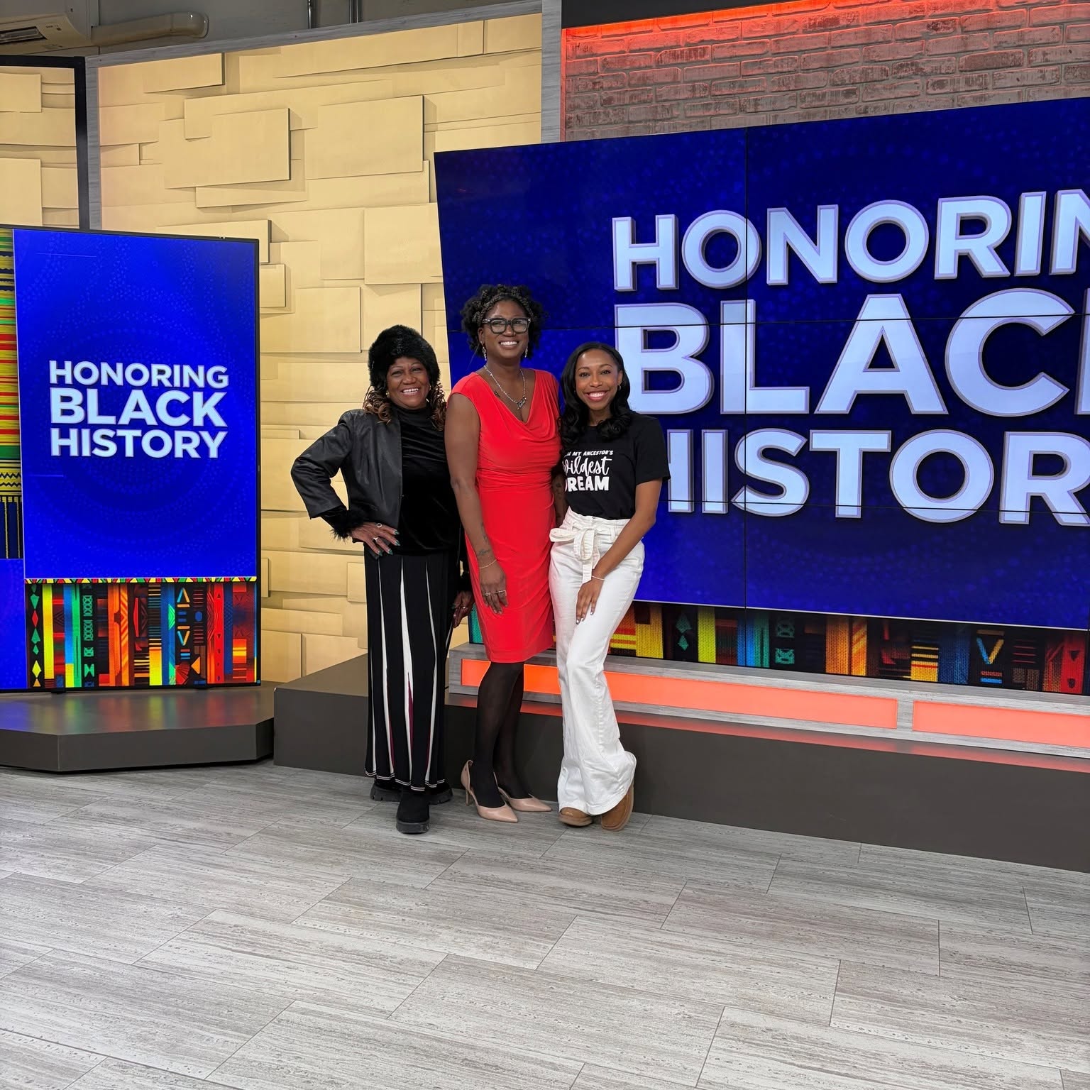 Three women standing together in front of a large screen with text honoring Black history, smiling and posing for the photo.