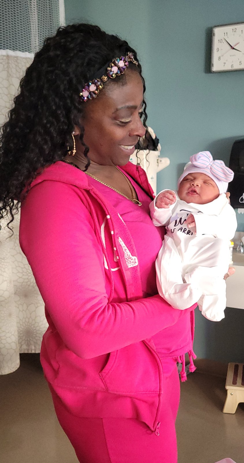 Woman in pink hoodie holding a newborn baby dressed in white with a pink hat, smiling indoors.