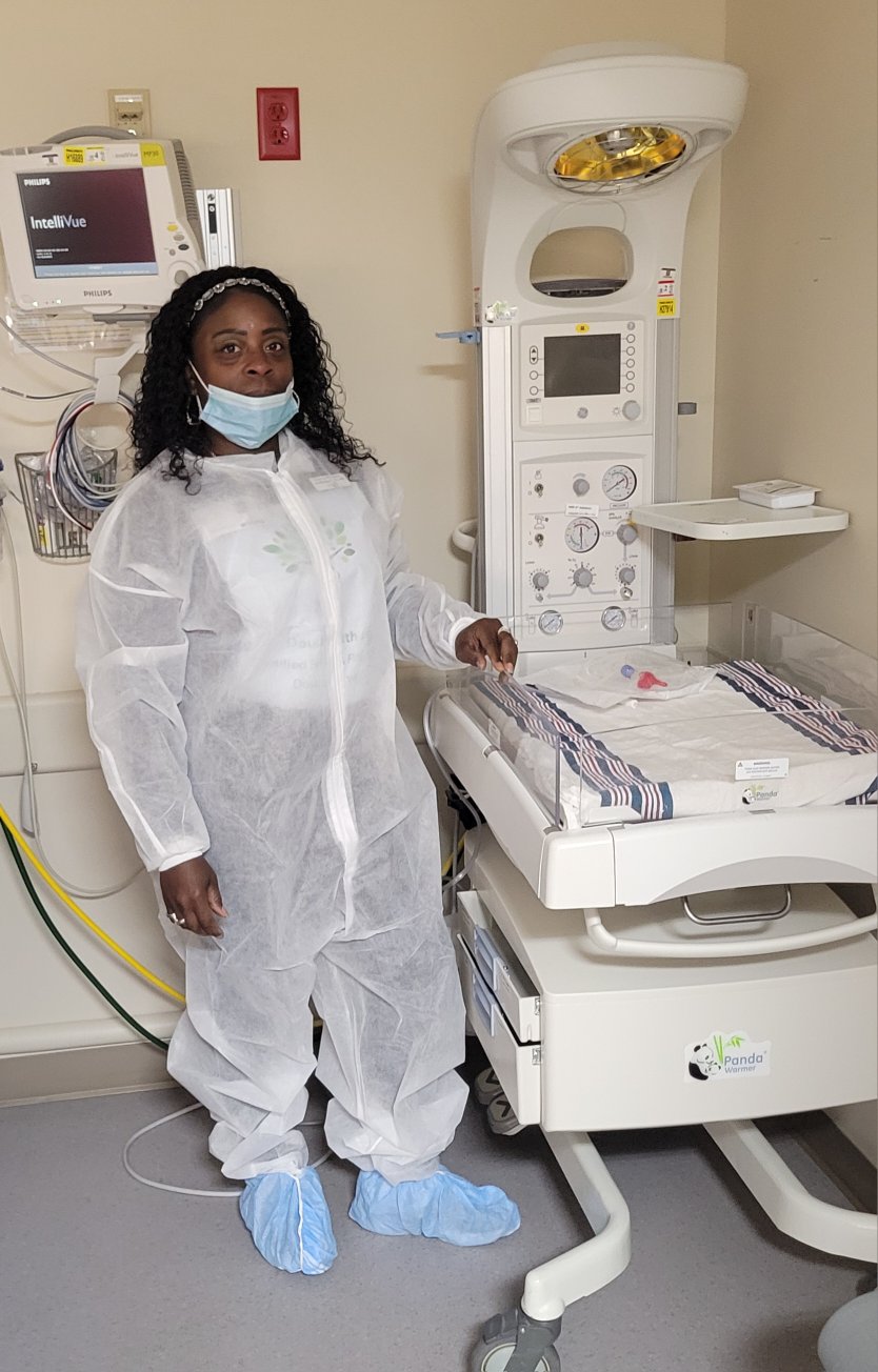 Person in protective gown, mask, and shoe covers standing next to medical equipment in a hospital room.