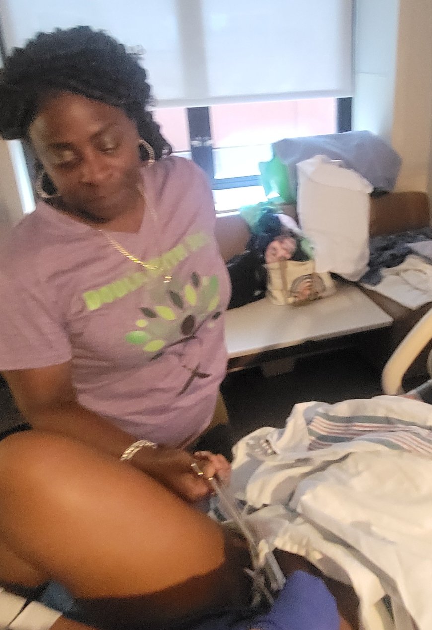 Woman standing next to a hospital bed with medical equipment and a patient lying down, in a room with a window.
