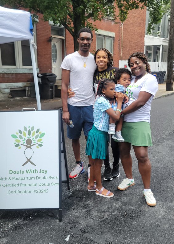 Group of five people standing outdoors on a street, smiling, with a sign about Doula services, trees, and buildings in the background.