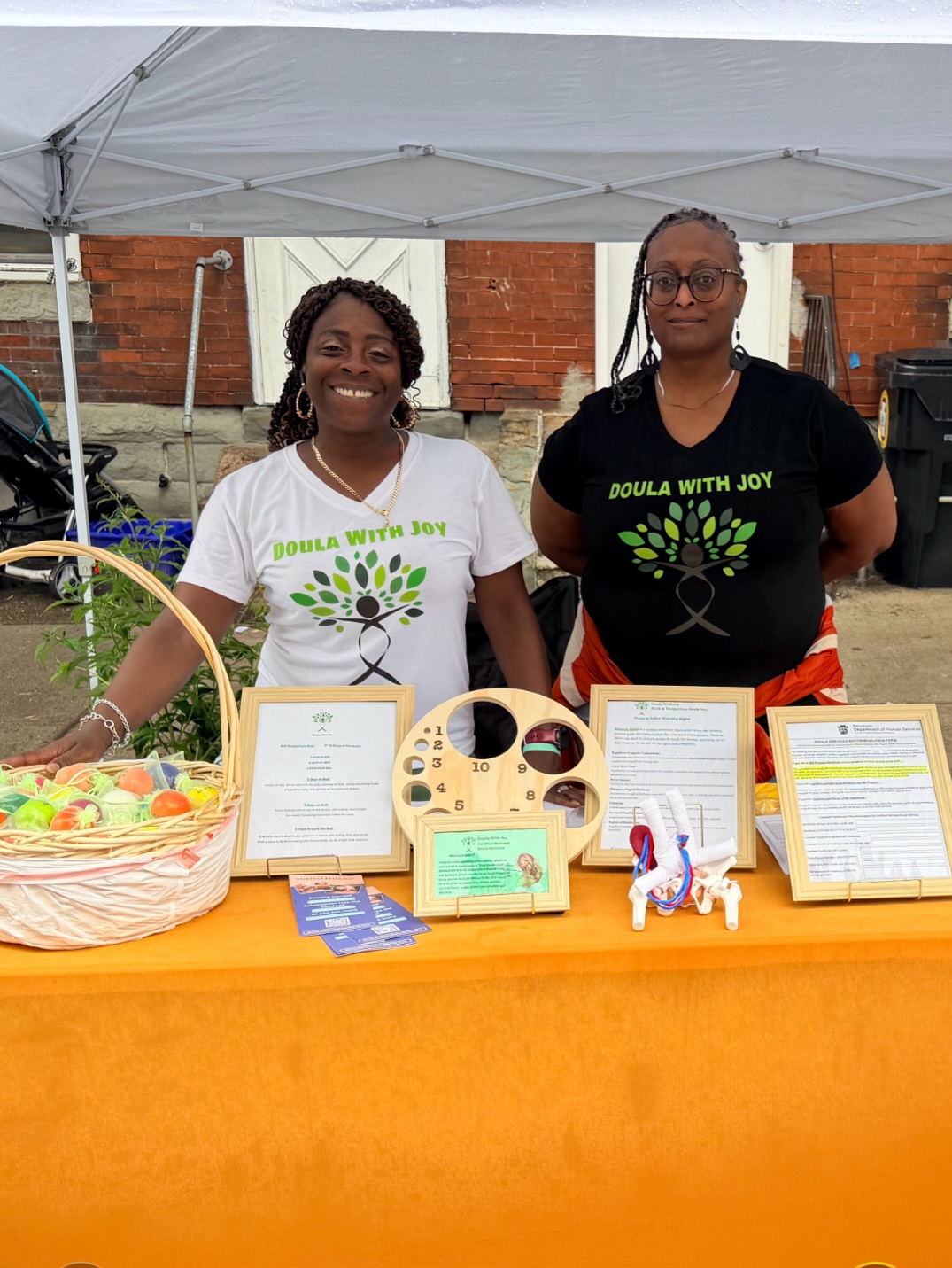 Two women stand behind a table with informational materials and a basket, under a white canopy outdoors, brick wall background.