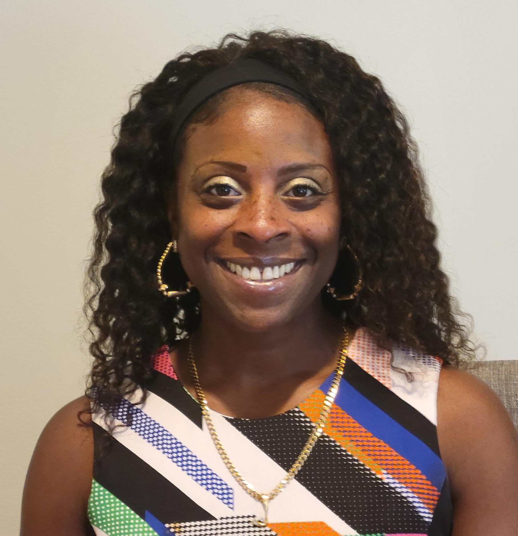 Smiling woman with curly hair wearing a colorful sleeveless top and gold jewelry.