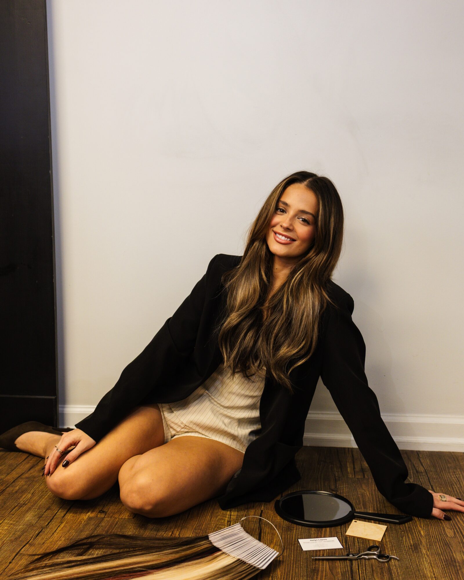Young woman with long hair sitting on wooden floor, smiling, wearing a black jacket and light-colored dress, against a plain wall.