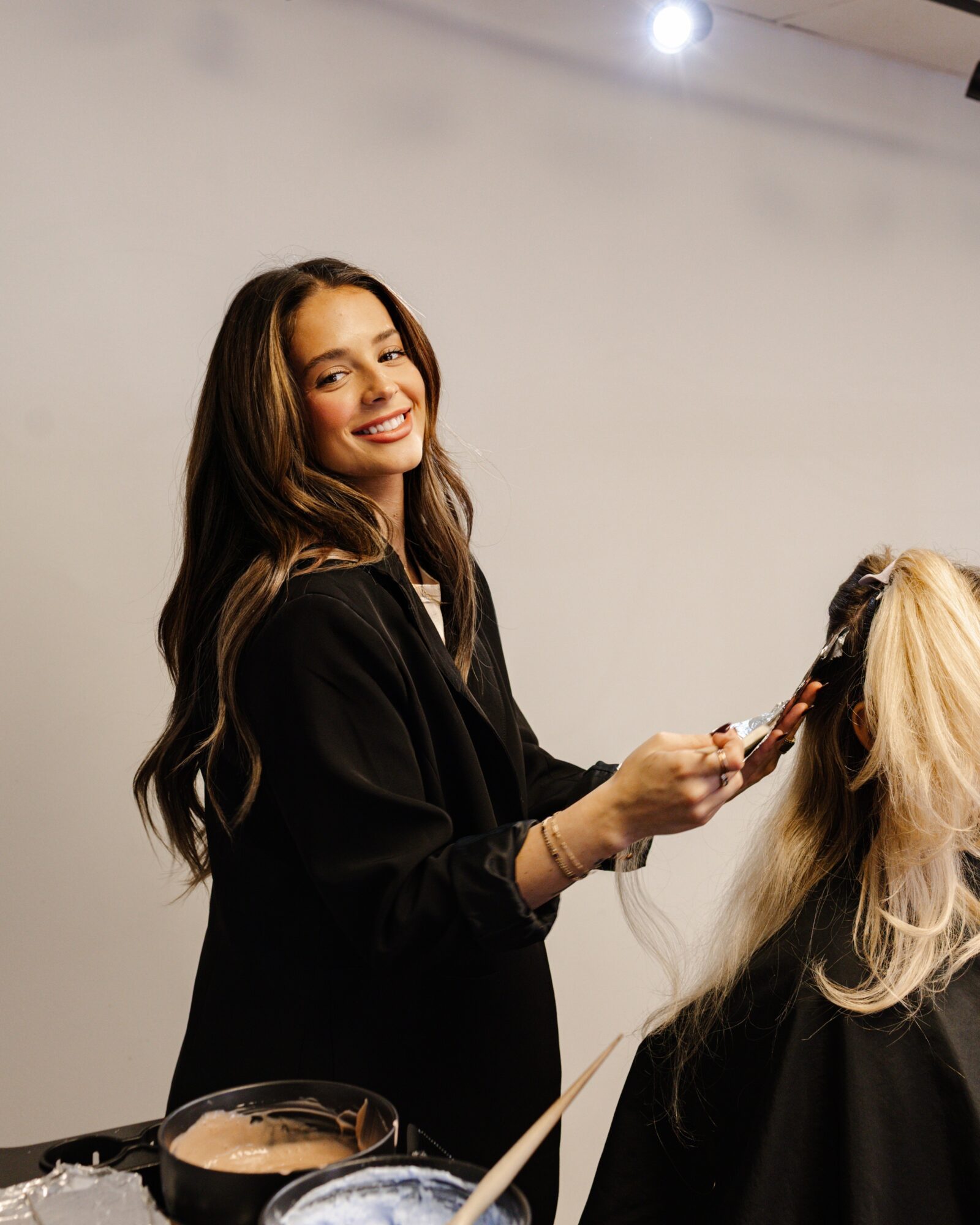 Woman smiling while styling a person's long blonde hair in a salon, with hair tools on a table nearby.