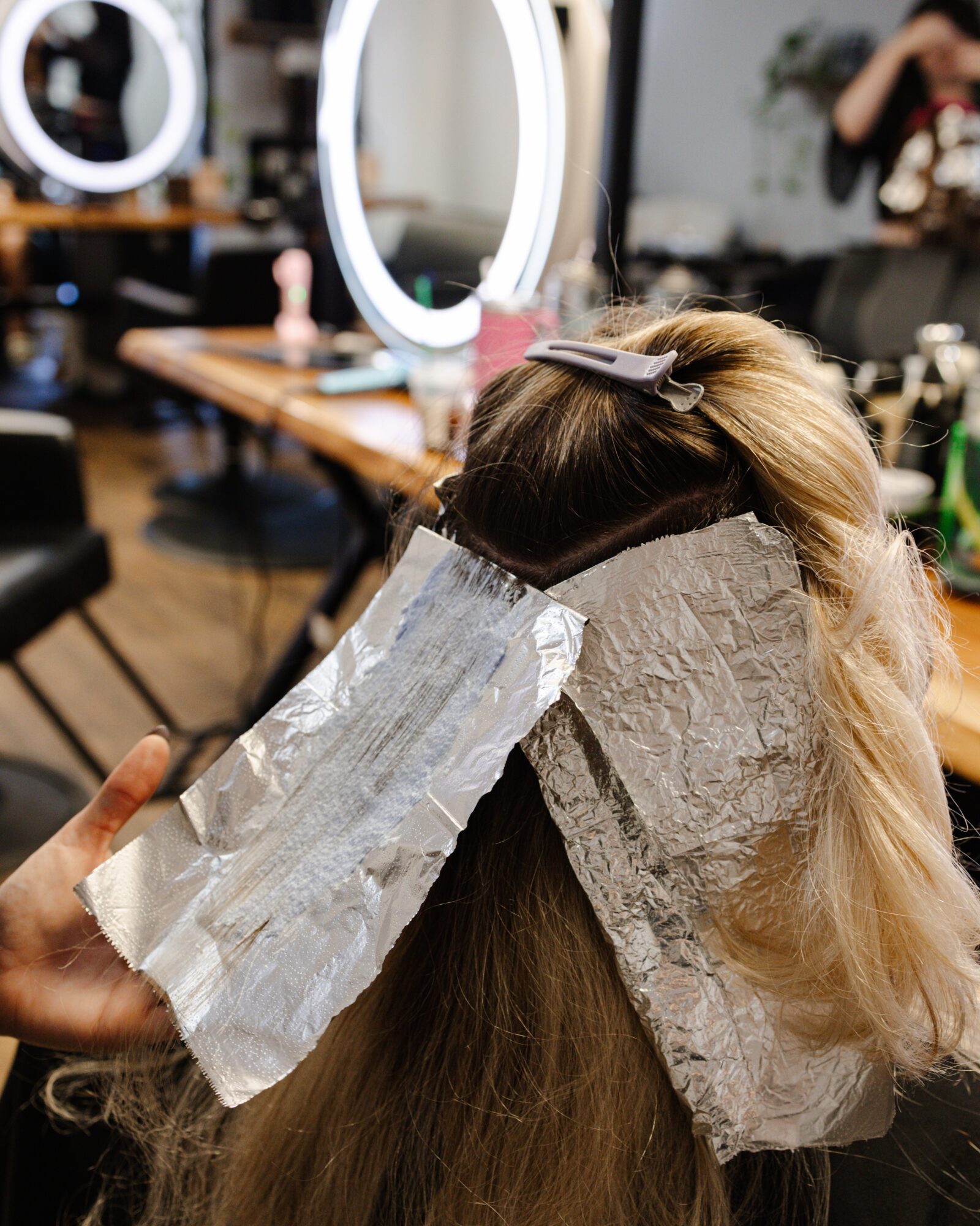 Person with hair covered in aluminum foil, sitting in a salon with mirrors and styling tools visible.