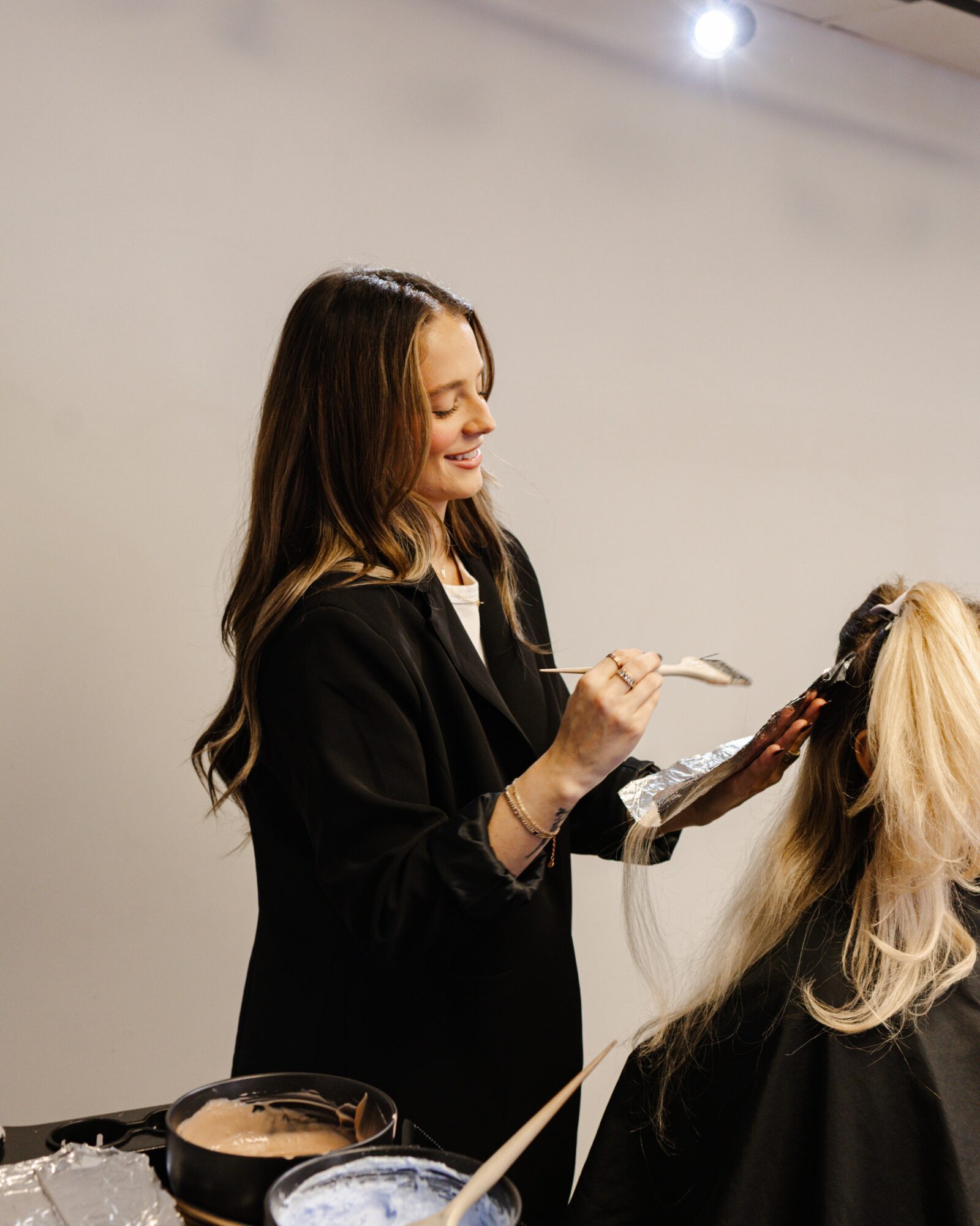 Woman with long hair styling another person's hair in a salon, smiling, with hairdressing tools nearby.