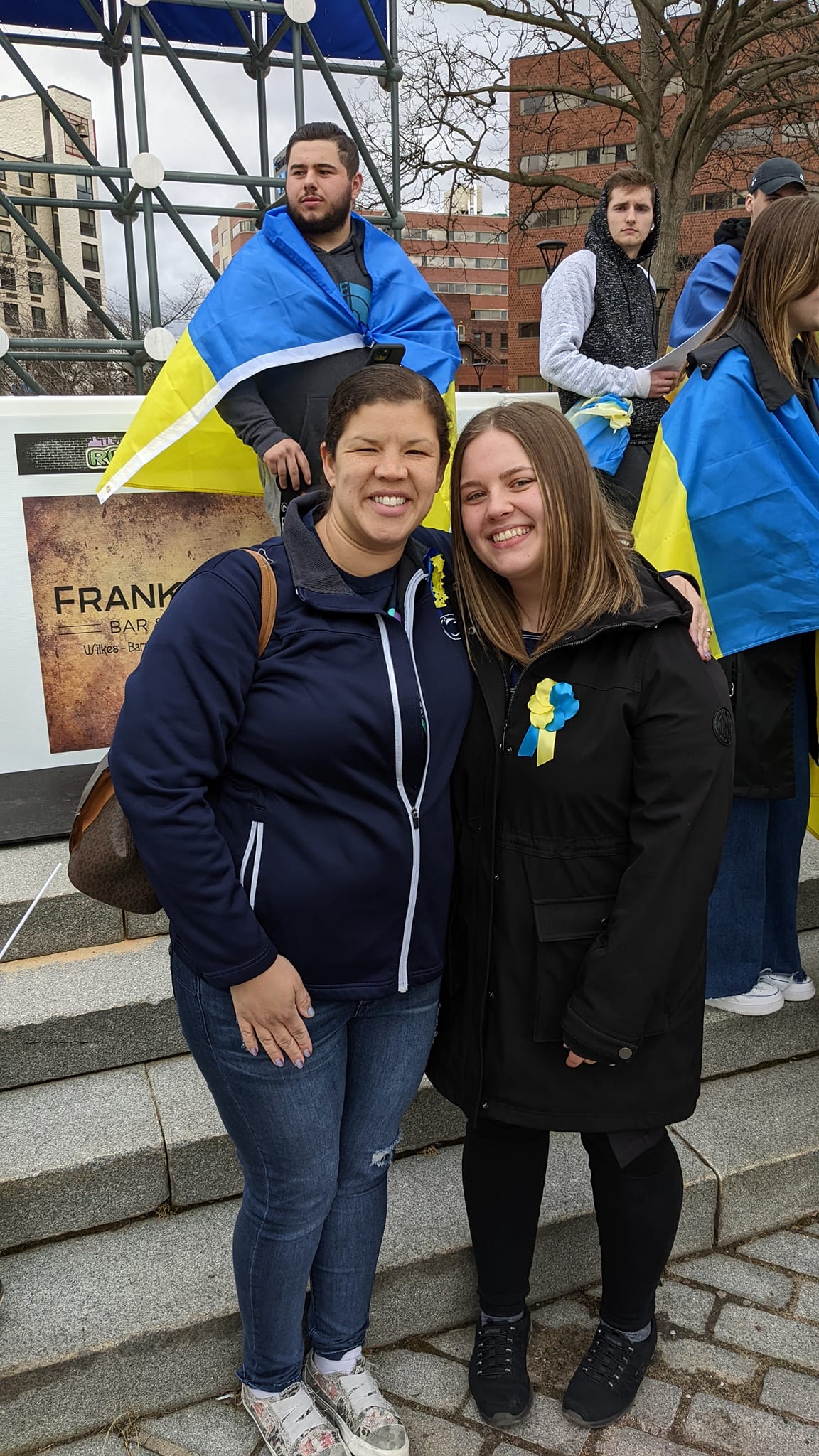 Two women smiling and standing together outdoors, with people and a Ukrainian flag in the background.