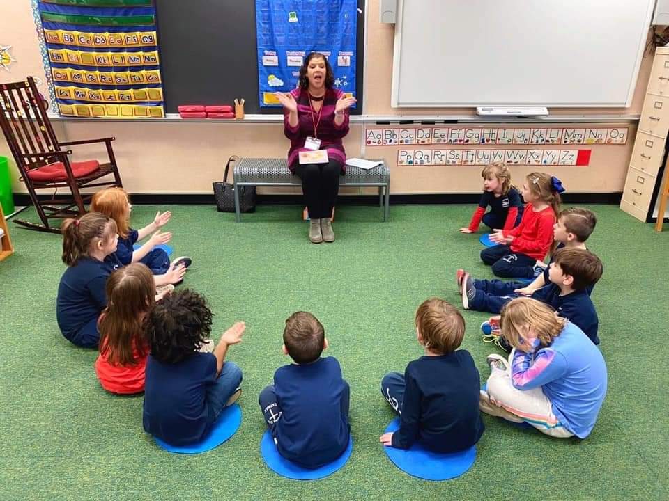 Teacher sitting on a bench in front of children sitting on the floor in a classroom.