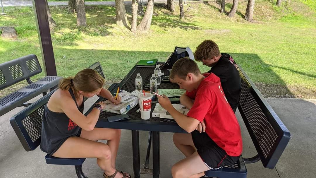 Three children sitting at a table outdoors, working on papers with drinks and supplies, surrounded by trees and grass.