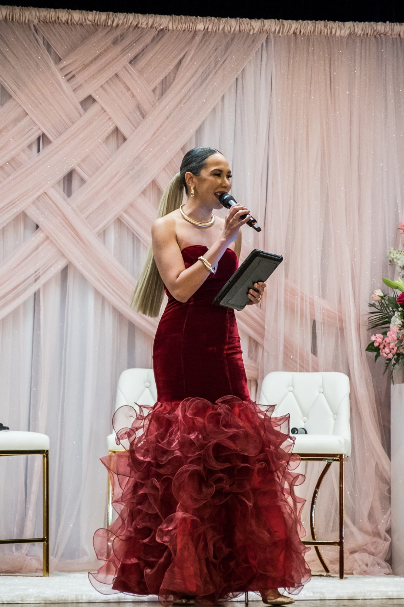 Woman in a red gown speaking into a microphone on stage with pink and white curtains and floral arrangements behind her.