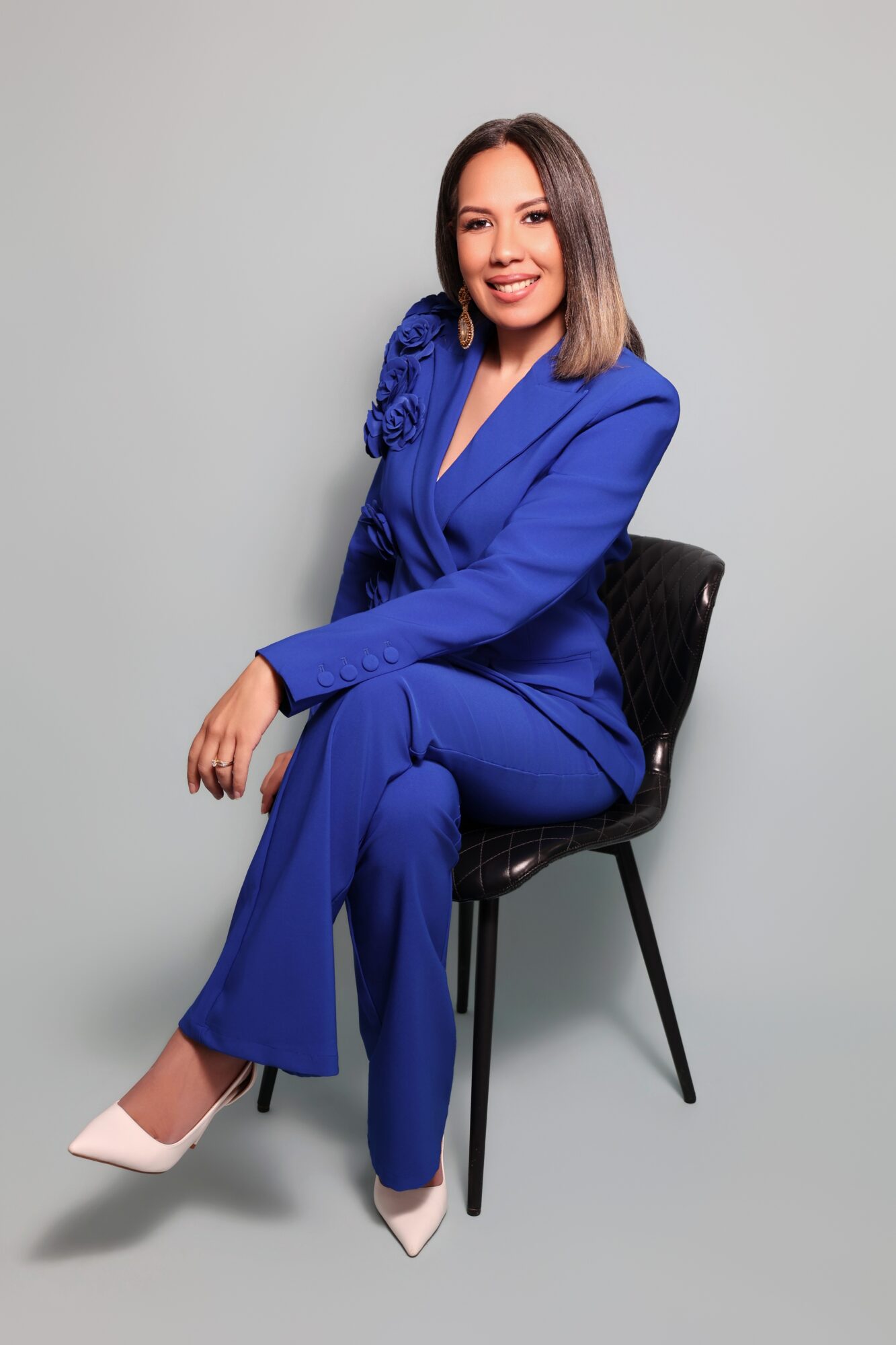 Woman in a blue suit sitting on a black chair, smiling, against a plain light gray background.
