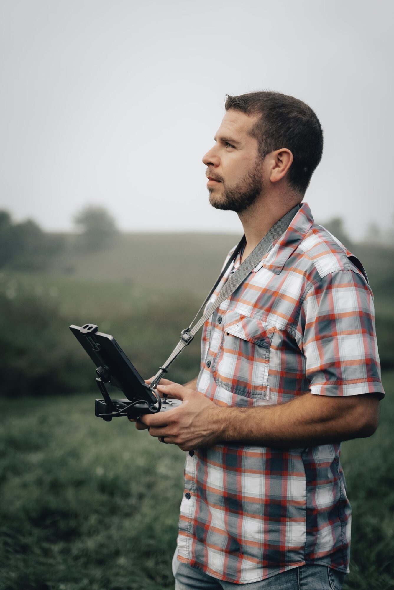 Man with a drone controller outdoors, wearing a plaid shirt, looking to the side, with a blurred natural background.