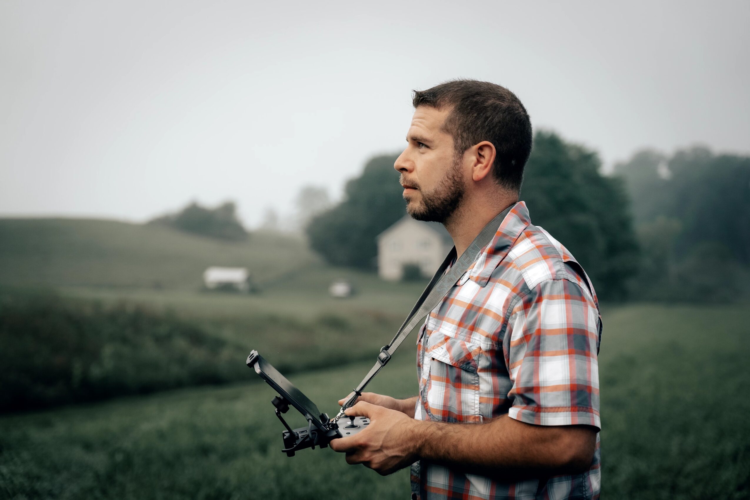 Man with a beard wearing a plaid shirt holding a drone controller outdoors in a grassy field.