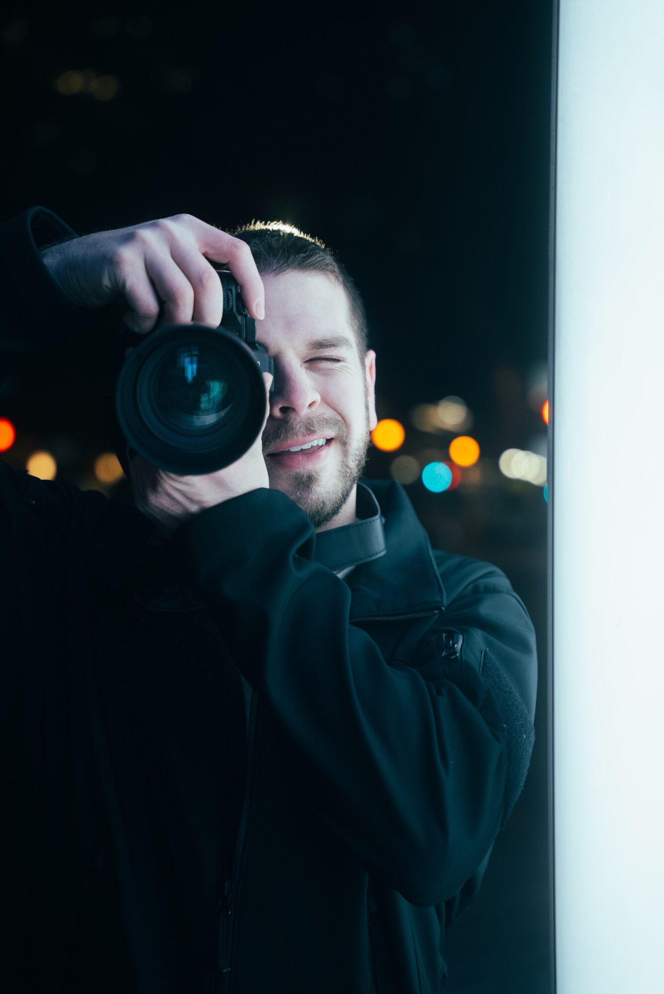 Person taking a photo with a camera at night, smiling, with city lights in the background.