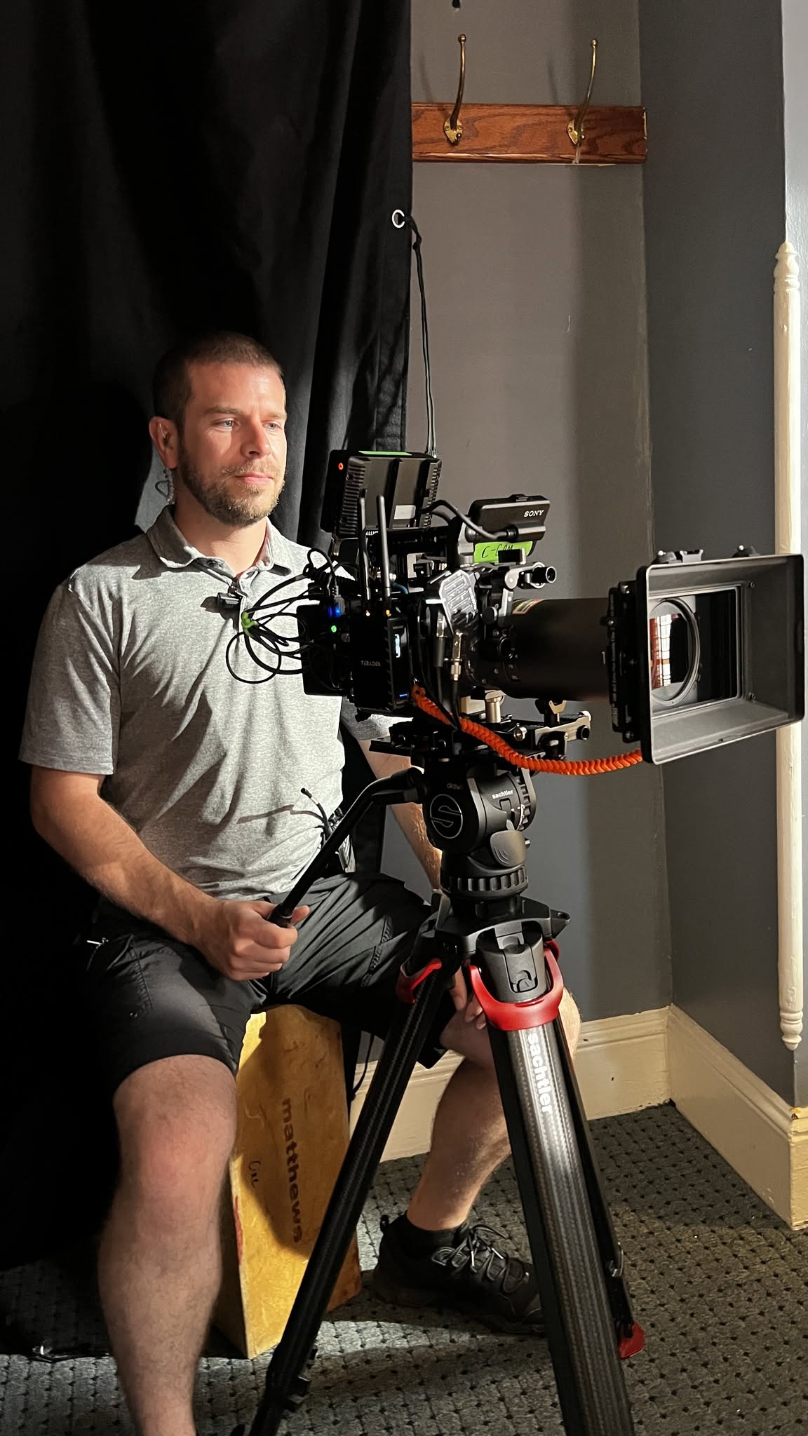Man operating professional video camera on tripod indoors, with black curtain and window nearby.