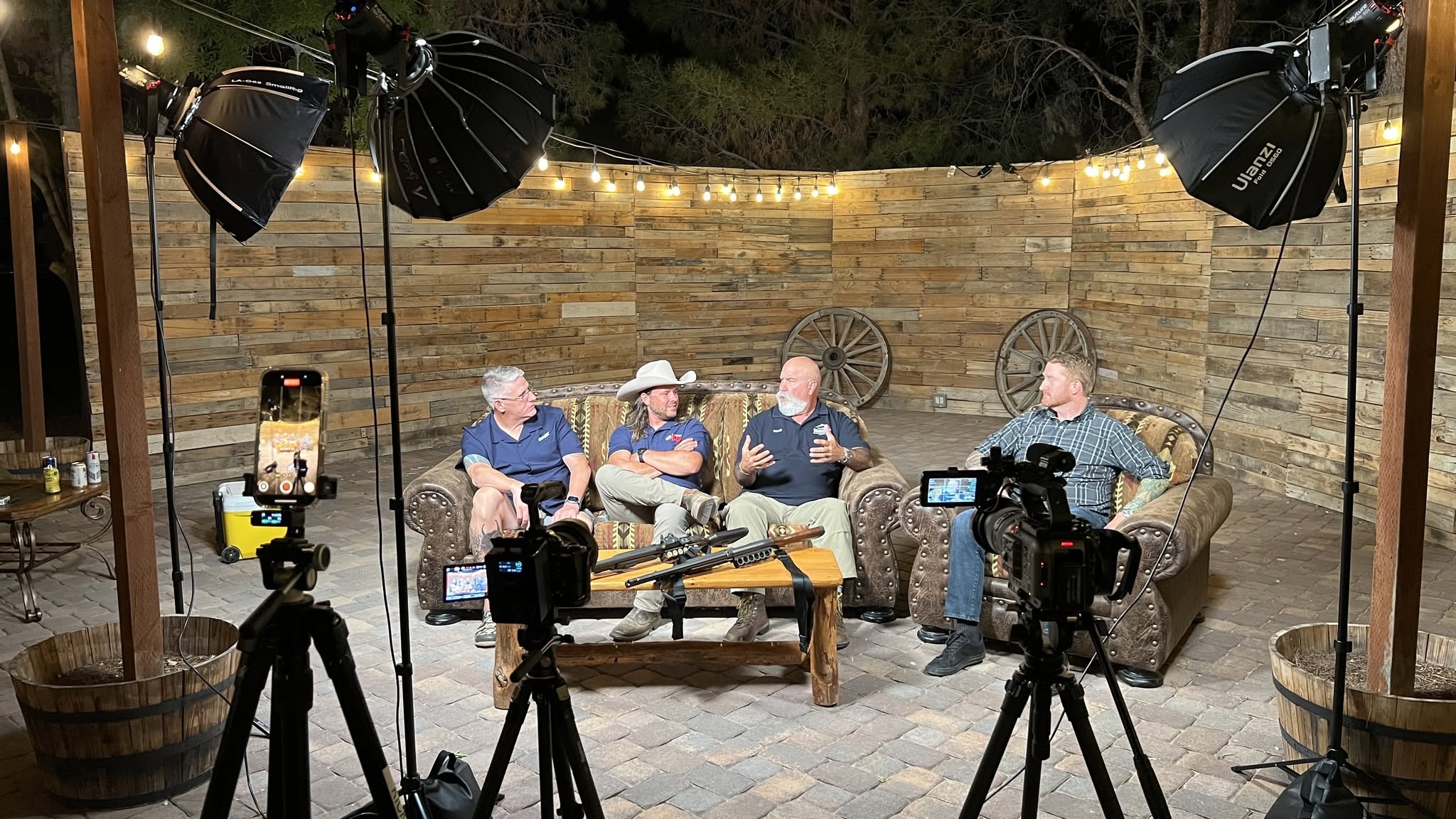 Four people sit on a sofa and chairs in an outdoor studio with lighting and cameras, against a brick wall background.