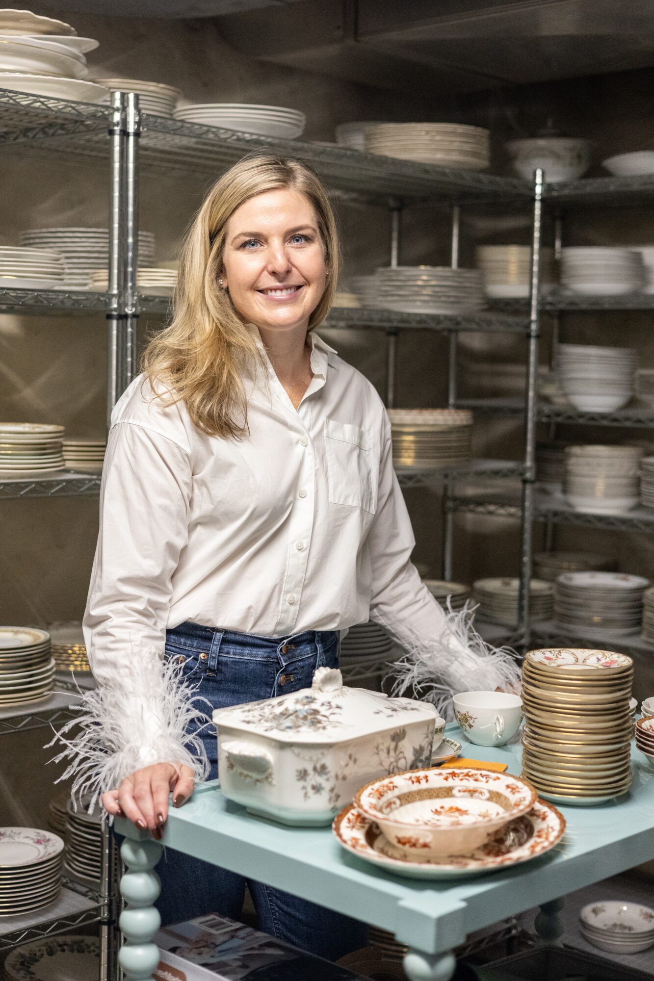 Woman with blonde hair in a white shirt stands in a storage room with shelves of plates and bowls, smiling at the camera.