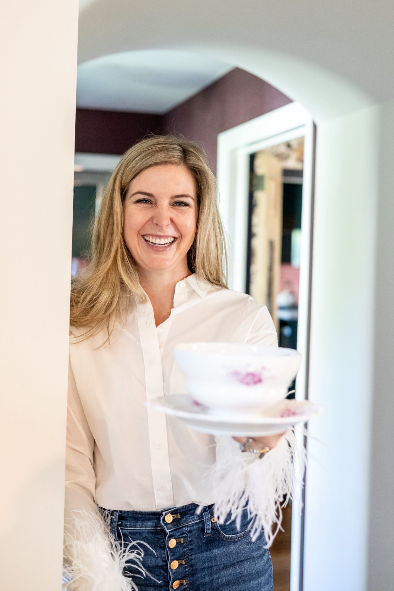 Woman with long blonde hair smiling, holding a cake on a plate, in a room with mirrors and a purple wall.