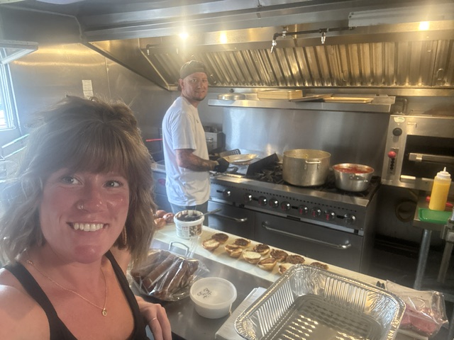 Smiling woman taking a selfie in a kitchen with a man cooking in the background, various kitchen items visible.