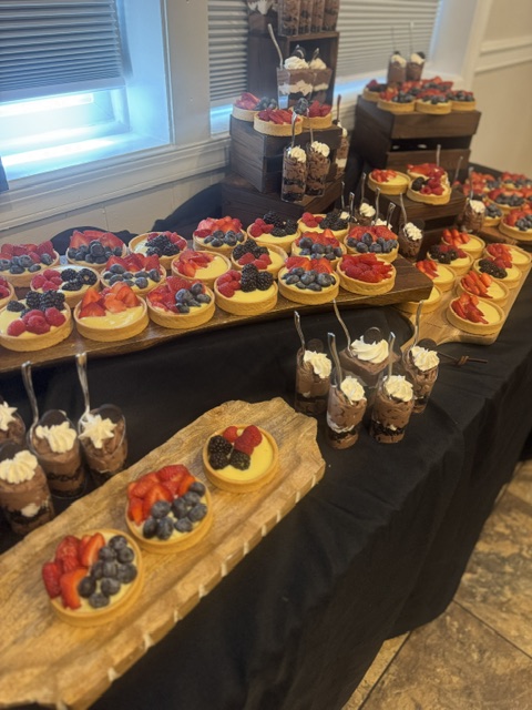 Dessert table with cupcakes, cake pops, and layered cakes on black tablecloth, near window with blinds, in a room with wooden floor.