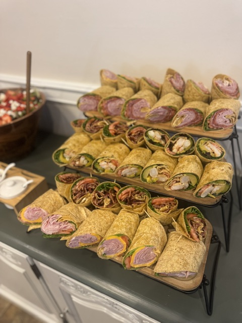 Tray of assorted wraps and pinwheel sandwiches on a black table, with a bowl of salad in the background.