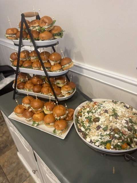 Three-tiered tray with mini burgers and onions, next to a large bowl of salad on a dark table against a white wall.