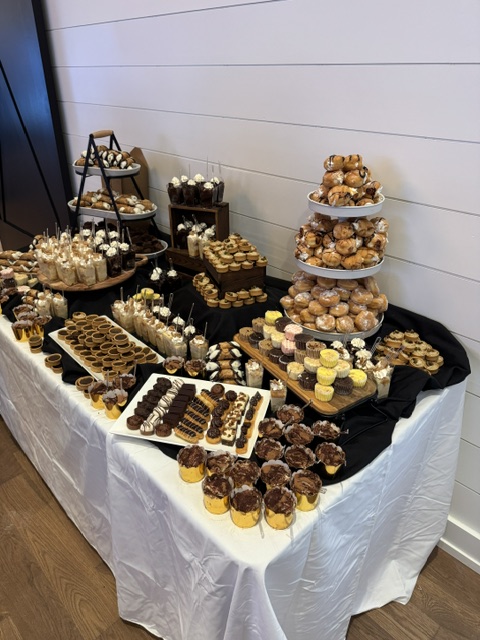 Table with assorted desserts and chocolates, some on trays and plates, on a white tablecloth, against a white wall.