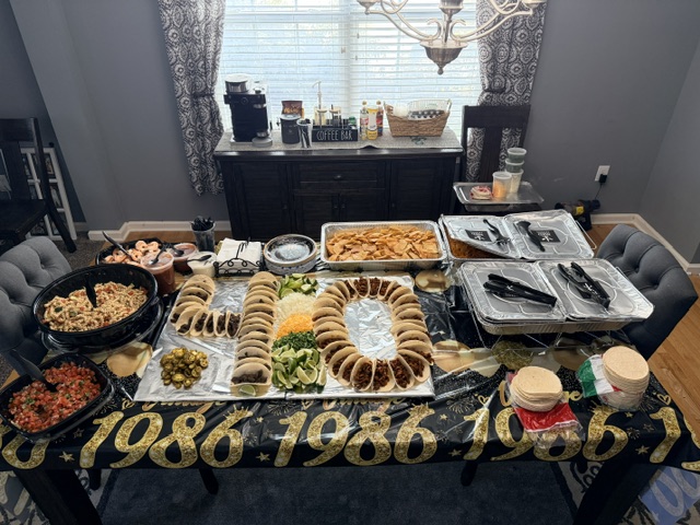 Table with various dishes, including pasta, bread, and salads, in a room with curtains and a chandelier.