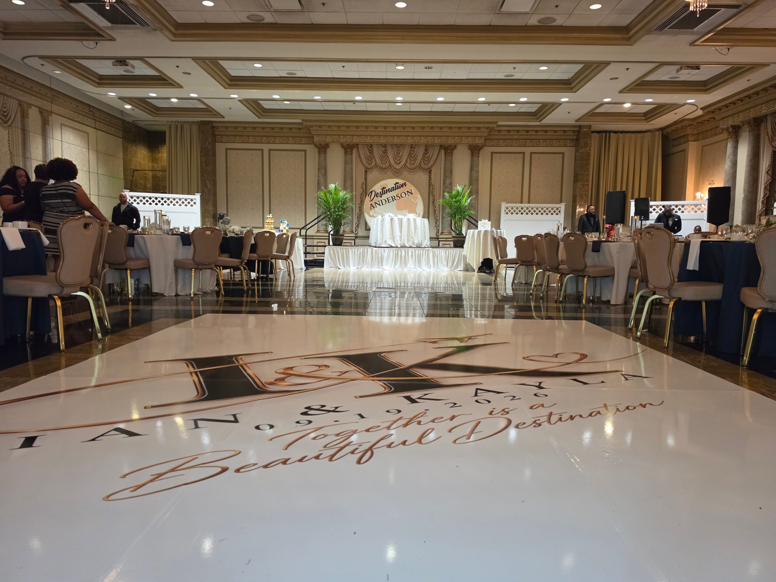 Elegant banquet hall with decorated tables, a stage with a draped backdrop, and a polished floor with writing, in a wedding setting.